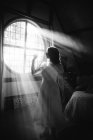 Vue latérale en noir et blanc de la femelle méconnaissable en robe debout contre fenêtre ronde dans la maison le jour ensoleillé — Photo de stock