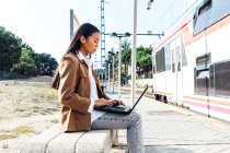 Орієнтована азіатська жінка в масці сидить на лавці на залізничній станції і переглядає ноутбук під час очікування поїзда. — стокове фото
