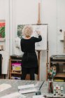 Обратный вид анонимной художницы, создающей рисунок человека карандашом, стоя у мольберта в студии — стоковое фото