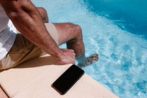 From above of crop unrecognizable male freelancer sitting at poolside near smartphone with black screen during remote work in summer — Stock Photo