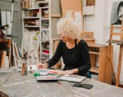 Сосредоточенная художница сидит за столом и рисует акварелью на бумаге во время работы в творческой мастерской — стоковое фото