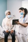 Young female doctor in medical uniform and stethoscope wearing face mask speaking and showing result on tablet to African American mature woman patient during appointment in clinic — Stock Photo