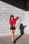 Junge tätowierte Frau in Activwear wirbelt Hula-Hoop-Reifen, während sie gegen Backsteinwände mit Schatten tanzt — Stockfoto