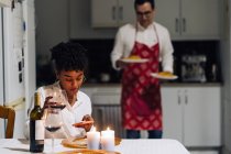 Черная женщина сидит за столом и просматривает смартфон, в то время как мужчина подает еду для романтического ужина дома — стоковое фото