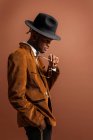 Vista laterale del giovane maschio afroamericano in abbigliamento alla moda e cappello guardando lontano su sfondo marrone — Foto stock