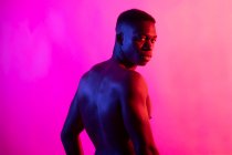 Серйозний молодий афроамериканець, спортсмен з голим тулубом, дивиться на фотоапарат на рожевому фоні в неоновій студії. — стокове фото