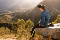 Vista lateral del viajero masculino en gafas VR interactuando con la realidad virtual mientras está sentado en la colina en terreno montañoso al atardecer - foto de stock