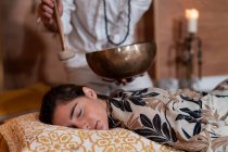 Cropped unrecognizable male spiritual therapist in beads playing Tibetan singing bowl over young female with closed eyes — Stock Photo