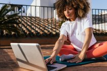 Mujer afroamericana enfocada con cabello rizado que elige un tutorial en línea en la computadora portátil mientras se sienta en la alfombra en la azotea y se prepara para la lección de yoga - foto de stock