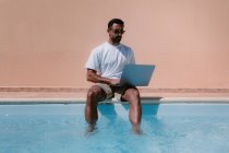 Männlicher Freiberufler mit Sonnenbrille sitzt am Pool und blättert im Netbook, während er in den Sommerferien aus der Ferne an einem Projekt arbeitet — Stockfoto