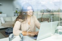 Attraverso la vista di vetro allegro giovane navigazione internet femminile su netbook a tavola con auricolare in appartamento — Foto stock