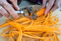 Растениеводство неузнаваемая женщина режет сырую морковь очистителем во время приготовления вегетарианской пищи в доме — стоковое фото