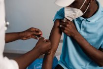 Médico negro en uniforme protector y guantes de látex vacunando a un paciente afroamericano irreconocible en la clínica durante el brote de coronavirus - foto de stock