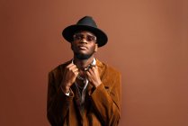 Junger afroamerikanischer Mann in trendiger Kleidung und Hut blickt in die Kamera auf braunem Hintergrund — Stockfoto
