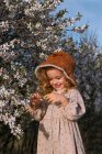 Adorabile bambino sorridente in abito in piedi vicino all'albero in fiore con fiori nel parco primaverile e guardando in basso — Foto stock