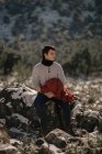 Jovem turista em roupas quentes contemplando a natureza enquanto sentado em pedra contra a montanha e olhando para longe à luz do sol — Fotografia de Stock