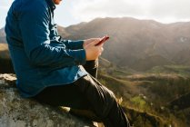 Side view of cropped unrecognizable tranquil male explorer with tablet sitting on rock and enjoying spectacular view of mountains on sunny day — Stock Photo
