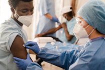 Seitenansicht einer Ärztin in Schutzuniform und Latexhandschuhen, die Arm mit Baumwolle und Alkohol desinfiziert, um männliche Afroamerikaner während des Coronavirus-Ausbruchs in der Klinik zu impfen — Stockfoto