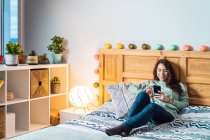 Молода щаслива жінка з бірюзовим светром і окулярами лежить на ліжку за допомогою мобільного телефону — стокове фото