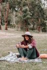Веселая женщина в шляпе сидит на одеяле на лугу в лесу и просматривает мобильный телефон, наслаждаясь пикником в Австралии — стоковое фото
