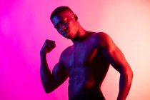 Fiducioso giovane atletico ragazzo nero con torso nudo guardando la fotocamera con le mani serrate in pugni in studio su sfondo rosa neon — Foto stock