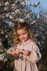 Adorable petit enfant souriant en robe debout près d'un arbre fleuri avec des fleurs au parc du printemps et regardant vers le bas — Photo de stock