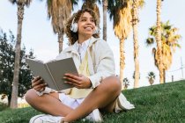 Angolo basso di felice femmina afroamericana in cuffia seduta in un parco esotico e leggere libro interessante in estate — Foto stock