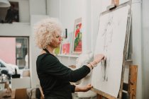 Female artist creating drawing of human with pencil while standing at easel in studio — Stock Photo