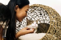 Side view of crop cheerful ethnic female caressing charming cat resting in wicker house — Stock Photo