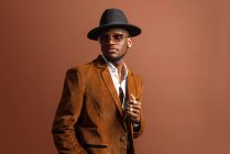 Junger afroamerikanischer Mann in trendiger Kleidung und Hut schaut vor braunem Hintergrund weg — Stockfoto