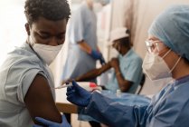 Medico donna in uniforme protettiva e guanti in lattice che vaccina il paziente afroamericano in clinica durante l'epidemia di coronavirus — Foto stock