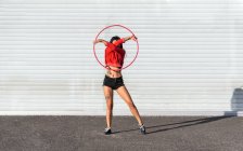 Junge tätowierte Frau in Activwear wirbelt Hula-Hoop-Reifen, tanzt mit Schatten gegen Ziegelwände und blickt im Sonnenlicht nach vorn — Stockfoto