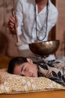 Проникливий нерозпізнаний чоловічий духовний терапевт у намистинах, який грає тибетську чашу для співу над молодою жінкою з закритими очима. — стокове фото