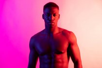 Joven atleta masculino afroamericano serio con el torso desnudo mirando la cámara en el fondo rosa en el estudio de neón - foto de stock