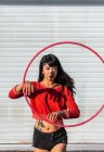 Young tattooed woman in activewear twirling hula hoop while dancing against brick walls with shadows — Stock Photo