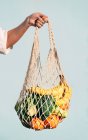Cultivado hembra irreconocible de pie con una variedad de frutas y verduras en bolsa de malla ecológica contra la pared azul en la ciudad - foto de stock