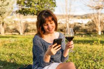 Donna spensierata seduta sul plaid sul prato e utilizzando lo smartphone mentre si gode il picnic nella giornata di sole in primavera — Foto stock