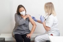 Spécialiste féminine en uniforme de protection et gants en latex vaccinant une patiente âgée en clinique pendant une éclosion de coronavirus — Photo de stock