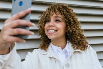 Ottimista donna afro-americana con acconciatura afro che si autoritratta sullo smartphone mentre si trova contro il muro metallico nell'area urbana della città — Foto stock