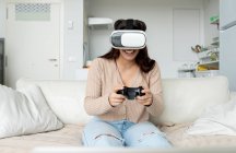 Unrecognizable cheerful female with gamepad experiencing virtual reality in goggles while playing video game on couch in house — Stock Photo