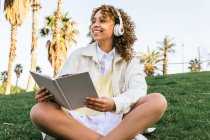 Bajo ángulo de la mujer afroamericana alegre en los auriculares sentados en el parque exótico y la lectura de libro interesante en verano - foto de stock