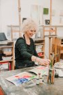 Konzentrierte Künstlerin sitzt am Tisch und malt mit Aquarellen auf Papier, während sie in der Kreativwerkstatt arbeitet — Stockfoto