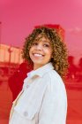 Mulher afro-americana encantada com penteado encaracolado em pé no fundo vermelho no estúdio olhando para longe — Fotografia de Stock