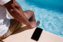 From above of crop unrecognizable male freelancer sitting at poolside near smartphone with black screen during remote work in summer — Stock Photo