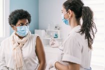 Especialista médica femenina en uniforme protector, guantes de látex y mascarilla facial vacunando a una paciente afroamericana madura en clínica durante el brote de coronavirus - foto de stock