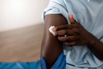 Especialista médico irreconocible en uniforme protector y guantes de látex que desinfecta el brazo con algodón con alcohol para vacunar al paciente afroamericano masculino en la clínica durante el brote de coronavirus - foto de stock