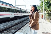 Side view of content ethnic female traveler standing with suitcase on platform at railroad station and talking on smartphone — Stock Photo