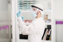 Вид збоку розмитого невідомого лікаря в захисній масці для обличчя та латексні рукавички з флаконом коронавірусної вакцини та шприцом, що стоїть у лікарняній кімнаті — стокове фото