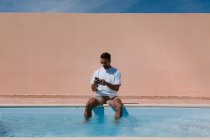 Serious male freelancer sitting at poolside with legs in water and speaking on mobile phone during remote work in summer — Stock Photo