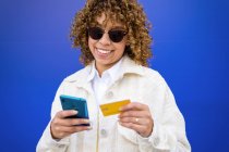 Контентна стильна афро-американська жінка, яка платить пластиковою карткою під час покупки через мобільний телефон, стоячи на синьому фоні в студії — стокове фото